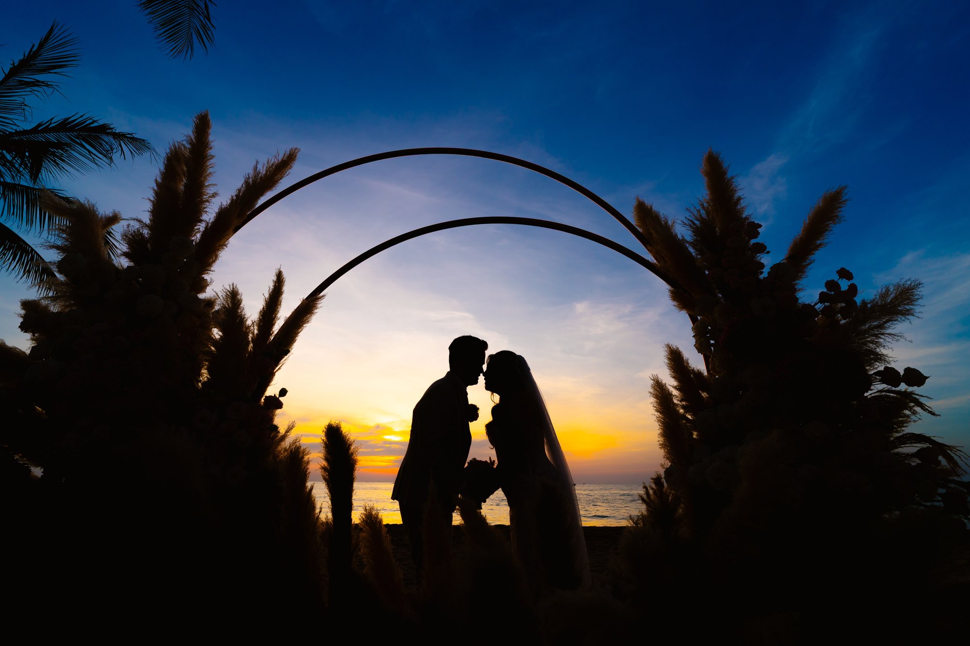 A couple kissing under a floral arch at sunset during a beach wedding at InterContinental Phu Quoc by TIMO Wedding